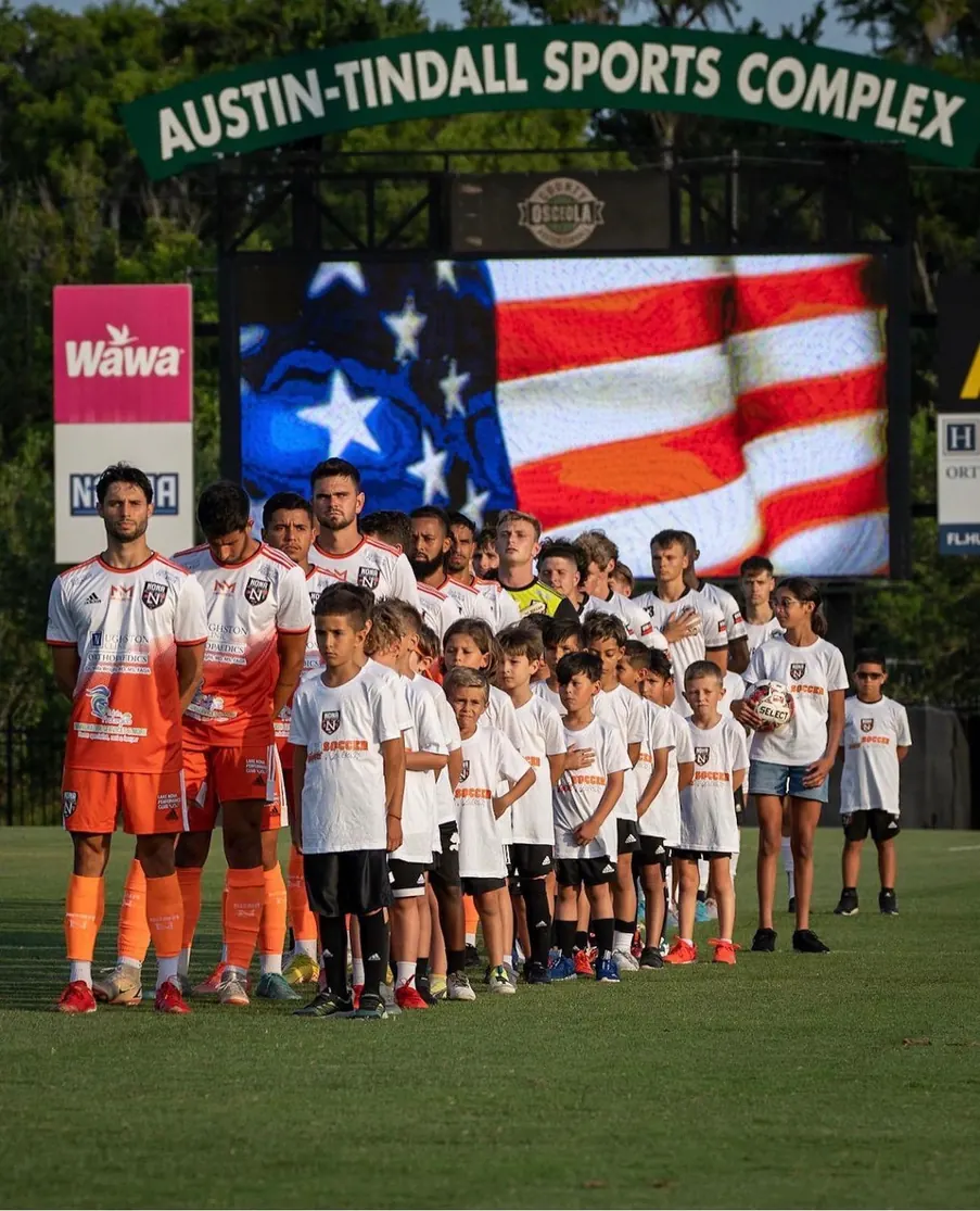 Elenco da Nona Soccer Academy perfilado em campo, diante de uma bandeira dos Estados Unidos exibida no telão do estádio