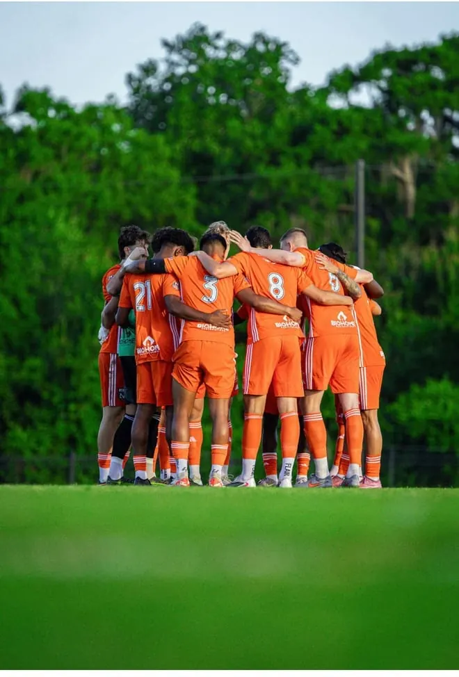 Atletas de uniforme laranja reunidos em roda no campo